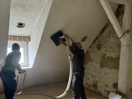 Zwei Handwerker arbeiten mit Einblasschläuchen an der Dachschräge eines Altbaus mit sichtbaren Holzbalken.