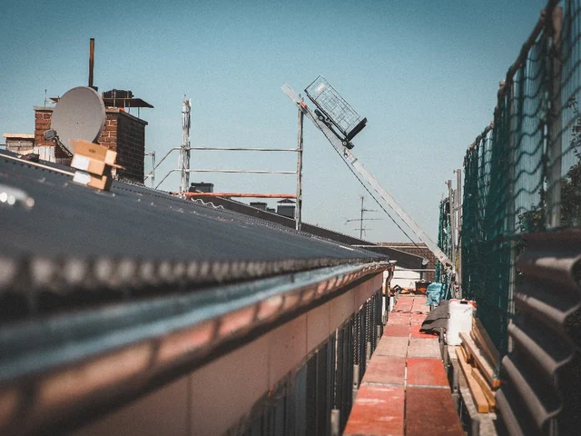 Dachfläche und Dachkante eines Bestandsgebäudes mit Schornsteinen und Arbeitsbühne im Hintergrund.
