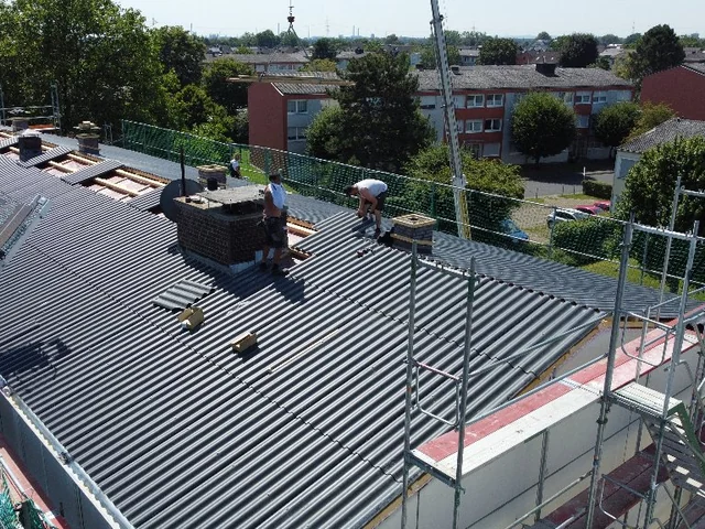 Dunkle Dachfläche eines Gewerbegebäudes mit Schornstein und umlaufendem Gerüst.