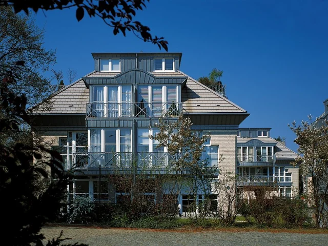 Mehrfamilienhaus mit dunklem Dach, mehreren Dachgauben und verglastem Vorbau.